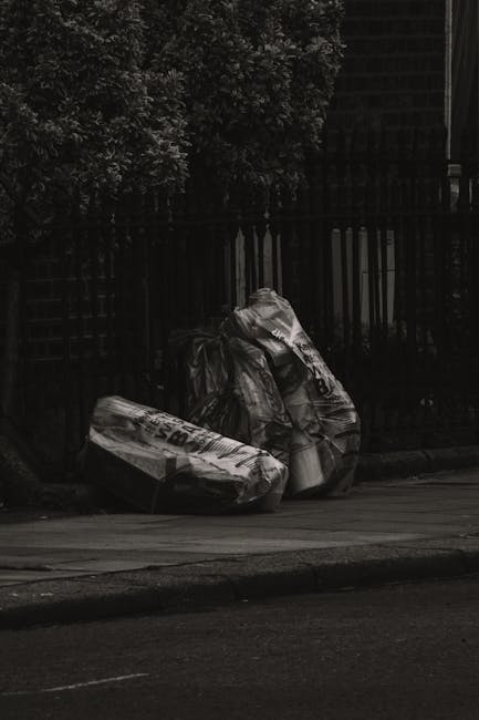 Two large black garbage bags filled with waste are placed on the pavement beside a wooden fence in an outdoor urban setting. The bags appear to be made of thick plastic, with some text printed on them