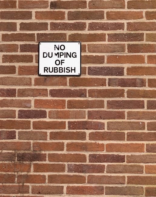 Two large black garbage bags filled with waste are placed on the pavement beside a wooden fence in an outdoor urban setting. The bags appear to be made of thick plastic, with some text printed on them